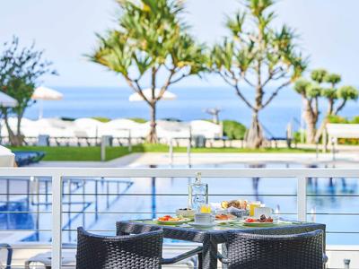 Terrasse mit Tisch und Stühlen, Blick auf Pool, grüne Bäume und blauen Himmel am Meer.