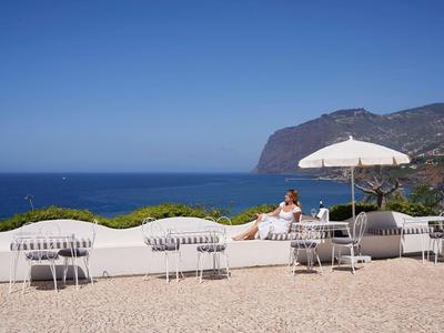 Terrasse mit weißen Sitzbänken, Tischen, Sonnenschirm und Aussicht auf das Blaue Meer und Berge.