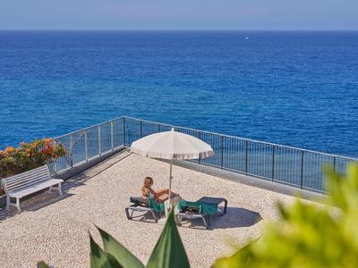 Eine Terrasse mit zwei Liegen, Sonnenschirm und Meerblick, umgeben von Geländer und Pflanzen.
