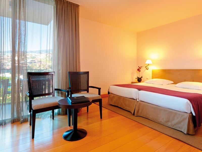 Camera d'hotel luminosa con letto matrimoniale, due sedie, tavolo e grande finestra con vista esterna.