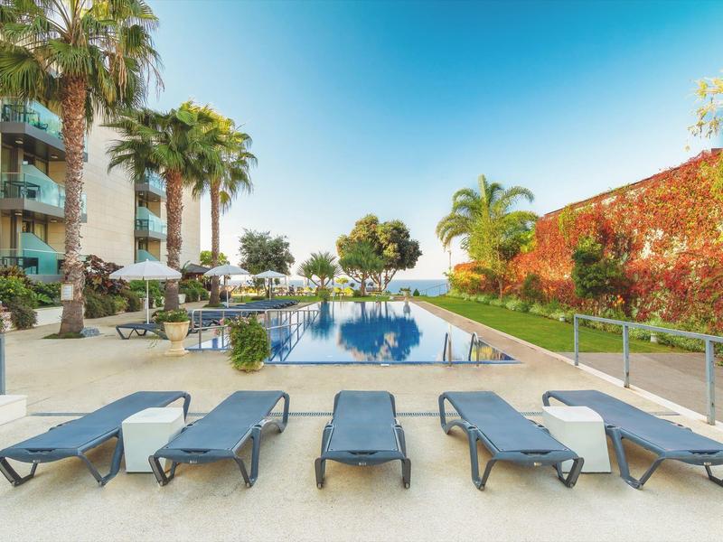Area piscina moderna con lettini, palme e cielo azzurro limpido in un hotel.