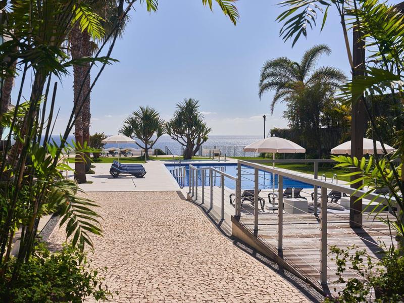 Piscina dell'hotel con lettini, ombrelloni e palme in un ambiente tropicale.