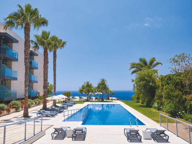 Piscina moderna dell'hotel con lettini, palme e vista sull'oceano sotto un cielo azzurro limpido.