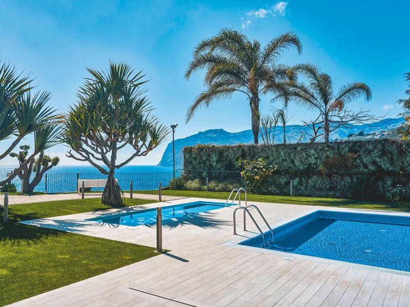 Piscina dell'hotel con prato, palme e vista mare sotto un cielo azzurro.