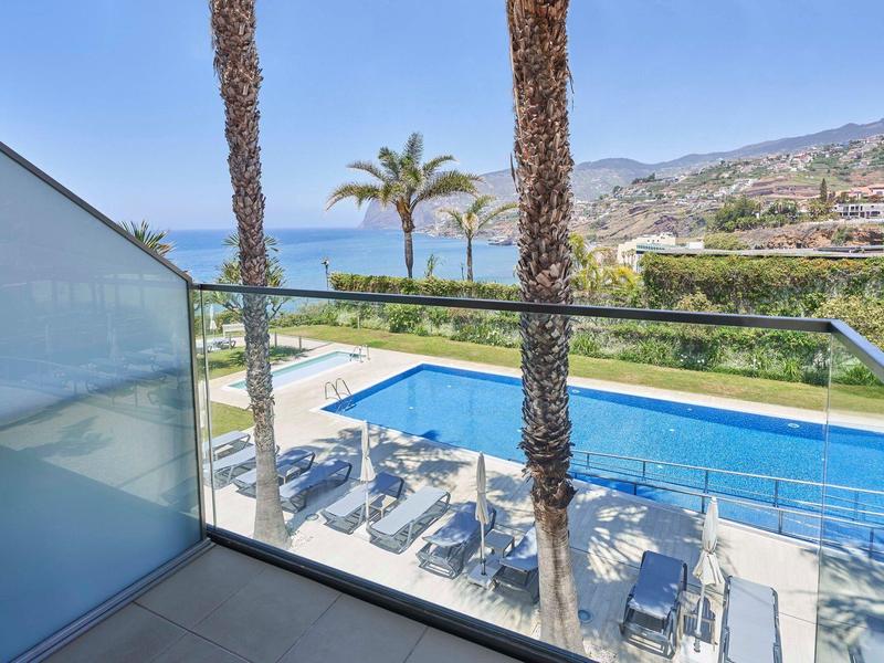 Vista da un balcone su una piscina con lettini, palme e il mare sullo sfondo.