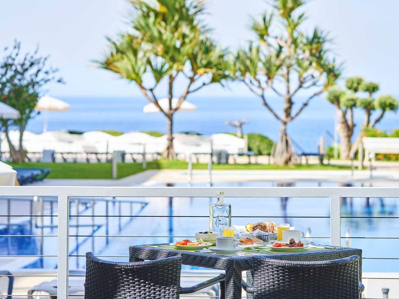 Tavolo con cibo davanti alla piscina, palme e spiaggia con tempo soleggiato.