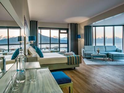 Camera d'hotel spaziosa con grandi finestre, letto, zona salotto e vista montagna al tramonto.