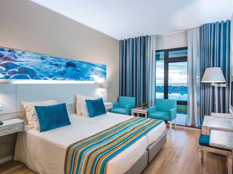 Camera d'hotel luminosa con due letti, accenti blu, balcone e vista sull'oceano.