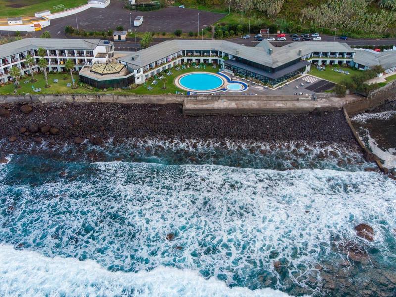 Hotel costiero con piscina, terrazza e onde che si infrangono su costa rocciosa.