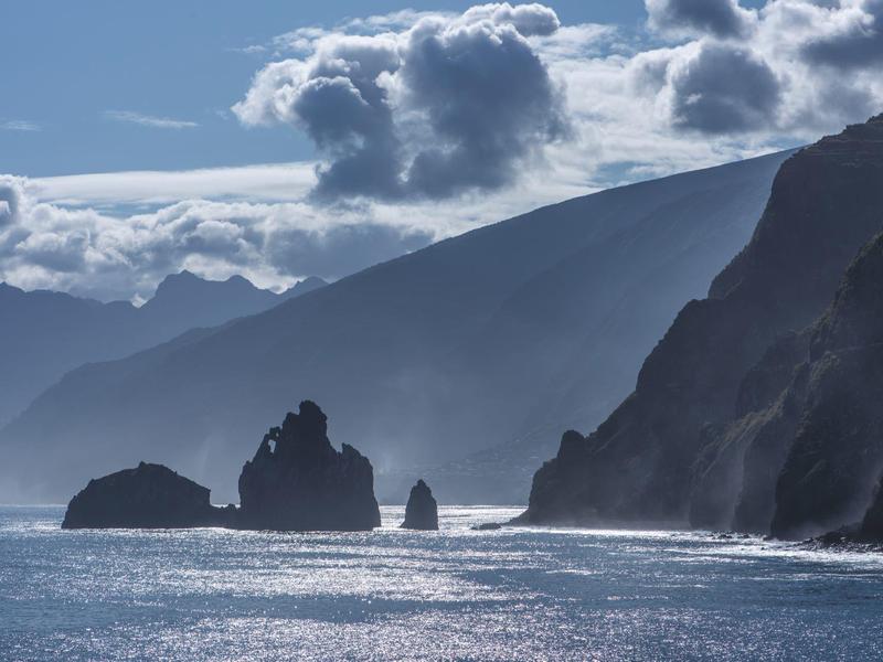 Paesaggio costiero drammatico con rocce nel mare e colline sotto un cielo nuvoloso.