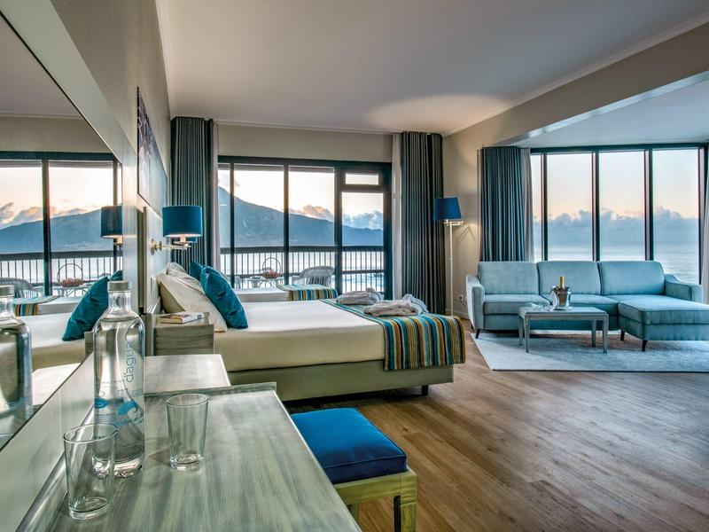 Camera d'hotel spaziosa con grandi finestre, letto, zona salotto e vista montagna al tramonto.