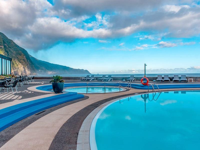 Piscina curva con acqua limpida che si affaccia su montagna e cielo nuvoloso in un resort sul mare.