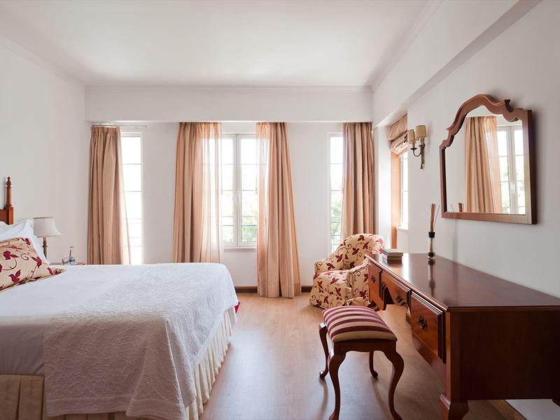 Camera d'hotel luminosa con letto matrimoniale, mobili classici e grandi finestre con tende.