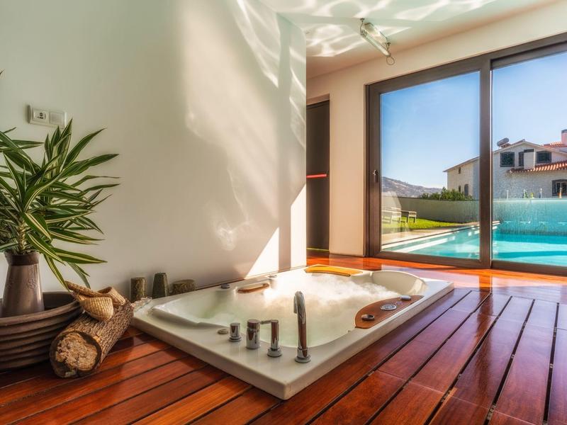 Area relax con jacuzzi luminoso, pavimento in legno e vista su piscina e case vicine.