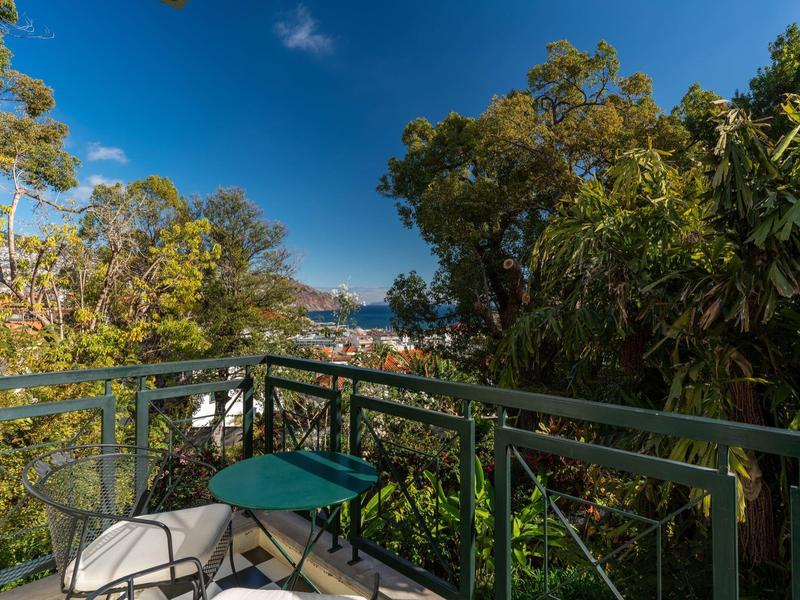 Balcone con sedie verdi e tavolo circondato da alberi, vista sul mare sullo sfondo.