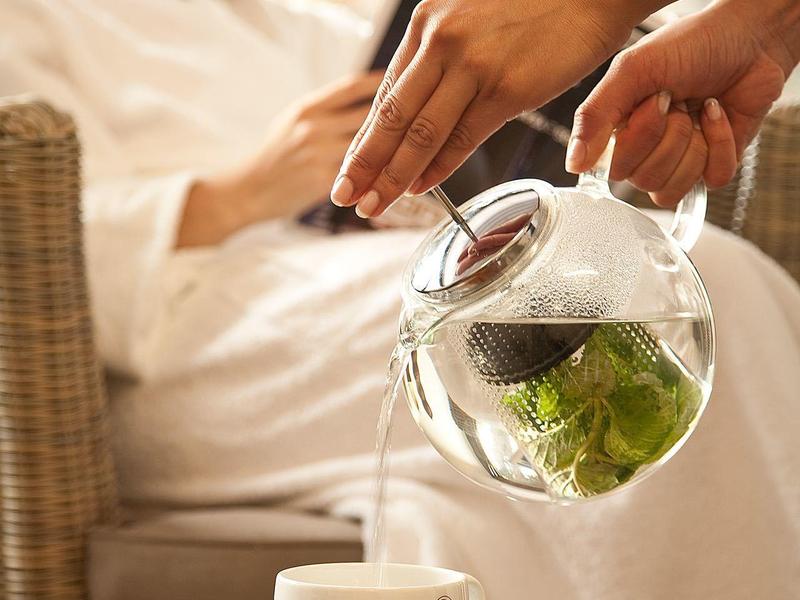 Una persona versa tè da una teiera di vetro in una tazza su un tavolo accanto a una donna rilassata in accappatoio.