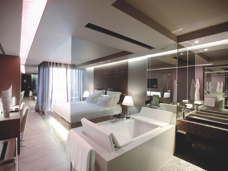 Chambre d'hôtel moderne avec lit, canapé, bureau et salle de bain avec cloison vitrée.