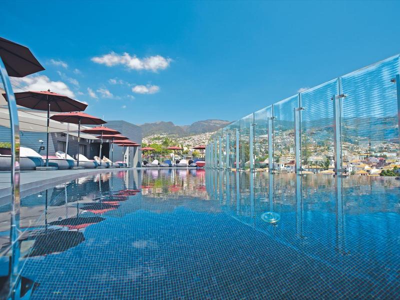 Piscine à débordement avec chaises longues et garde-corps en verre sous un ciel bleu avec des montagnes en arrière-plan