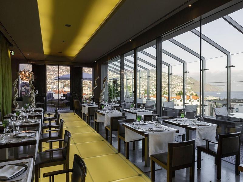 Restaurant élégant avec façade en verre et vue sur la mer, mobilier moderne avec tables blanches.