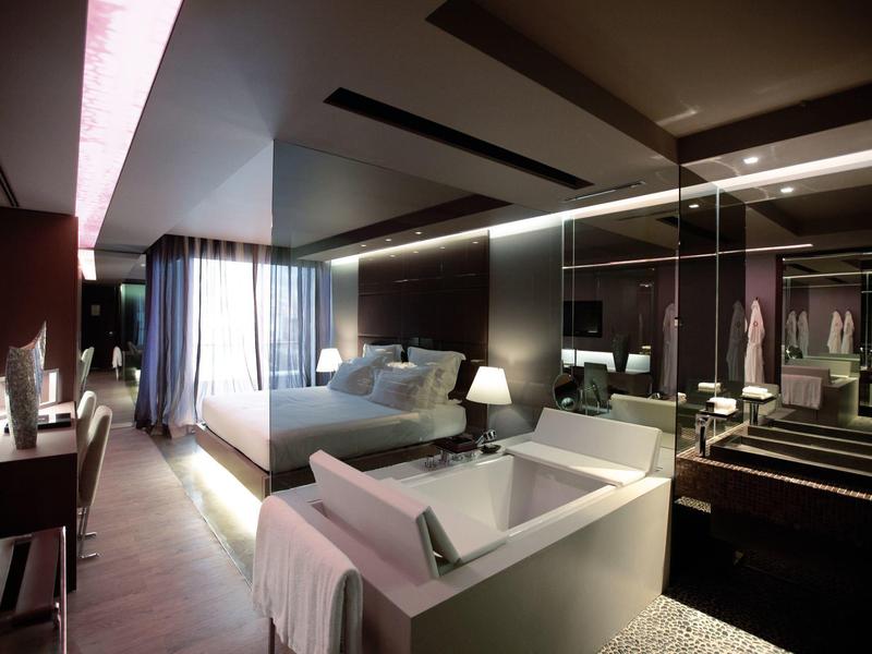 Chambre d'hôtel moderne avec lit confortable, canapé et grande paroi en verre donnant sur dressing.