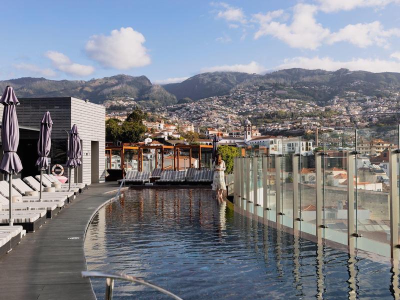 Piscine à débordement sur une terrasse sur le toit avec vue sur une ville et des montagnes sous un ciel partiellement nuageux