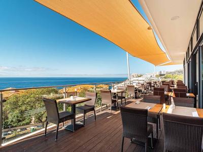 Große Terrasse mit Tischen und Stühlen, Blick auf Meer und blauen Himmel, gelbe Markise oben.