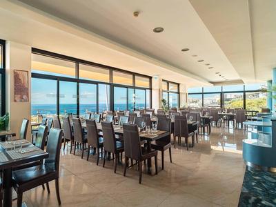 Großer Restaurantraum mit dunklen Holztischen und Stühlen, großen Fenstern und Meerblick.