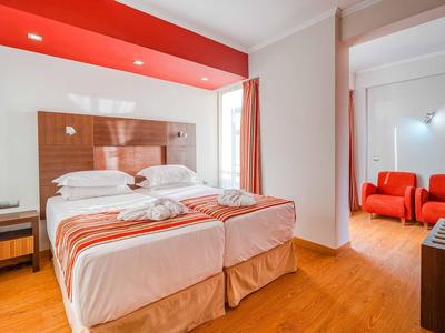 Helles Zimmer mit zwei Betten, roter Wand, Holzboden und angrenzendem Sitzbereich mit roten Sesseln.