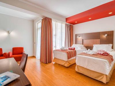 Helles Hotelzimmer mit zwei Einzelbetten, orangen Sesseln, Vorhängen und Holzboden.