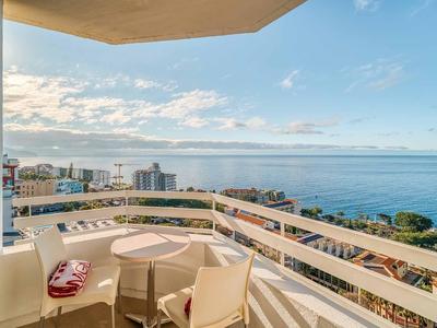 Balkon mit Tisch und Stühlen, Blick auf Stadt und Meer unter blauem Himmel mit Wolken.