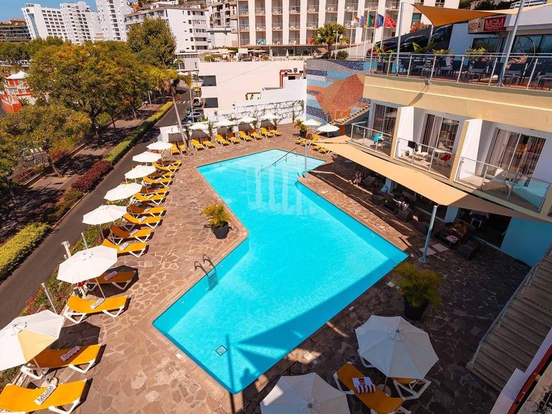 Großer blauer Pool mit gelben Liegestühlen und weißen Sonnenschirmen umgeben von Gebäuden und Bäumen.