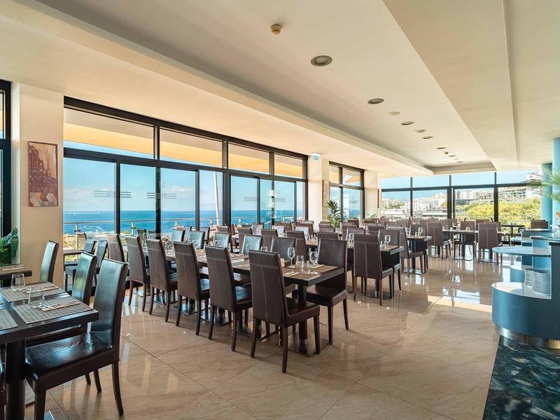 Großer Restaurantraum mit dunklen Holztischen und Stühlen, großen Fenstern und Meerblick.