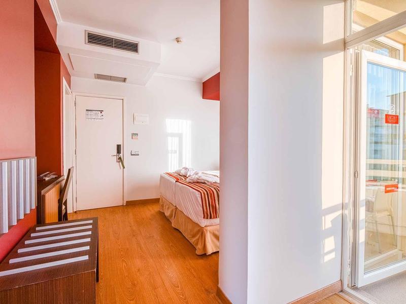 Helles kleines Zimmer mit Holzfußboden, roter Wand, Bett und Zugang zu Balkon durch Glas-Schiebetür.