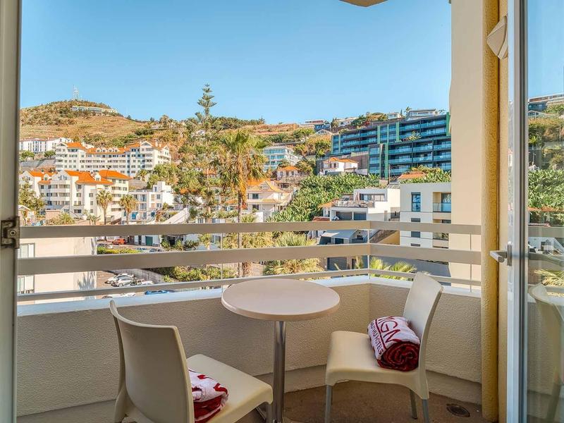 Balkon mit kleinem runden Tisch, zwei Stühlen und Blick auf bunte Häuser und Hügel im Hintergrund.