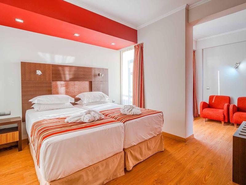 Helles Zimmer mit zwei Betten, roter Wand, Holzboden und angrenzendem Sitzbereich mit roten Sesseln.