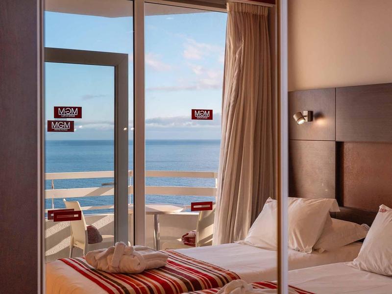 Helles Hotelzimmer mit Doppelbett, Meerblick durch Glasfenster, Balkon und blauem Himmel im Hintergrund.