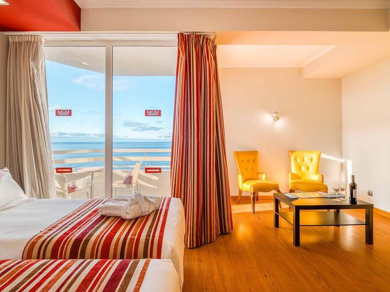 Helles Hotelzimmer mit zwei Betten, Meerblick, roten Vorhängen und gelben Sesseln neben einem Tisch.