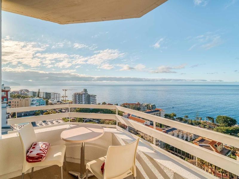 Balkon mit Tisch und Stühlen, Blick auf Stadt und Meer unter blauem Himmel mit Wolken.
