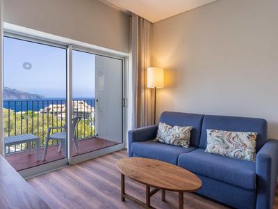 Wohnzimmer mit blauem Sofa, Holztisch, Lampe und Balkon mit Meerblick bei Tag.