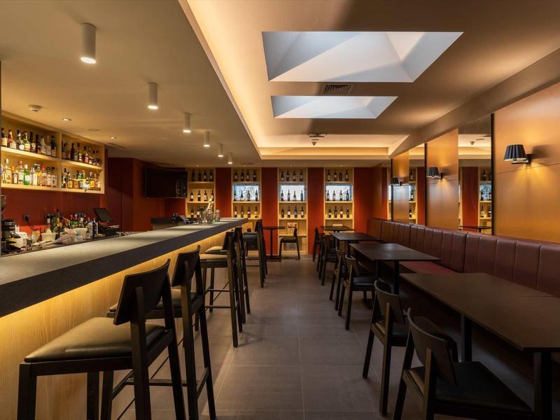 Moderne Bar mit langer Theke, Hockern, Deckenbeleuchtung und großen Fenstern an der Rückseite.