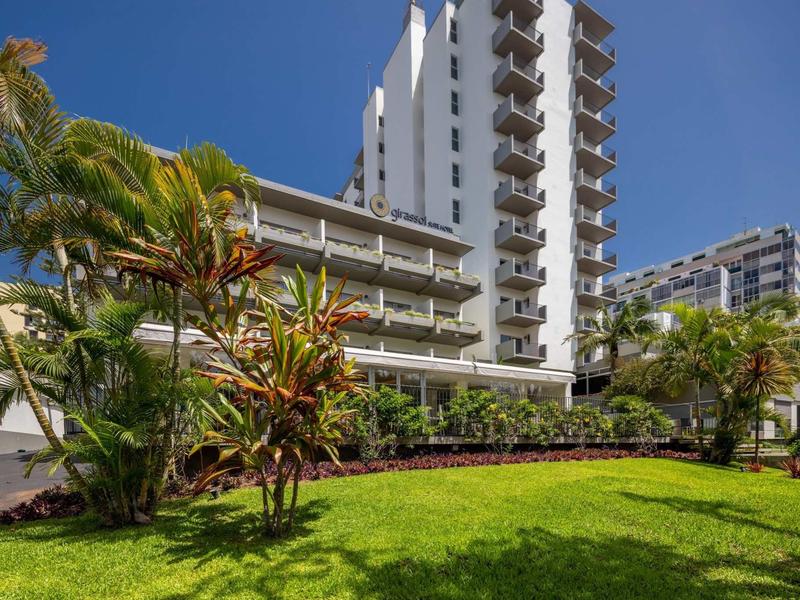 Hohes weißes Wohngebäude mit Balkonen, davor grüner Rasen und tropische Pflanzen unter blauem Himmel.
