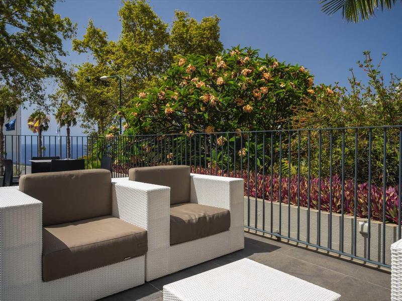 Zwei moderne weiße Sessel mit braunen Kissen auf Balkon mit Pflanzen und Meerblick bei klarem Himmel.