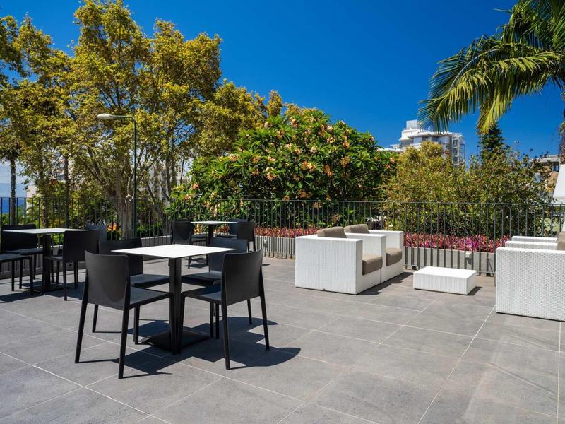 Moderner Außenbereich mit schwarzen Stühlen, weißen Sofas und grünen Bäumen unter blauem Himmel.