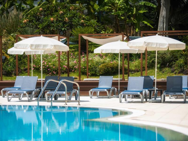 Blauer Pool mit weißen Sonnenschirmen und blauen Liegestühlen, umgeben von grünen Pflanzen.