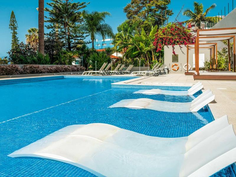 Großer Pool mit weißen Liegestühlen im Wasser und Palmen im Hintergrund bei klarem Himmel.