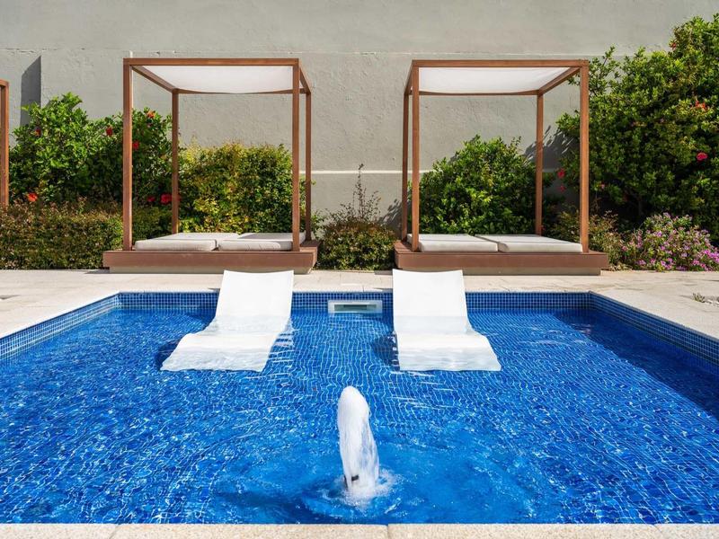 Blauer Pool mit Springbrunnen, zwei Liegestühlen und Pflanzen vor grauer Wand im Außenbereich.