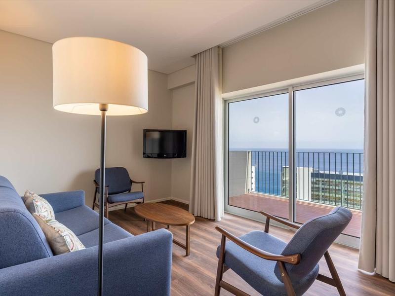 Helles Wohnzimmer mit blauen Sesseln, Holztisch, Stehlampe und großer Fensterfront zum Balkon.