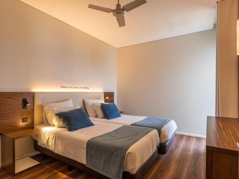 Modernes Schlafzimmer mit zwei Einzelbetten, Holzboden, Deckenventilator und schlichter Beleuchtung.