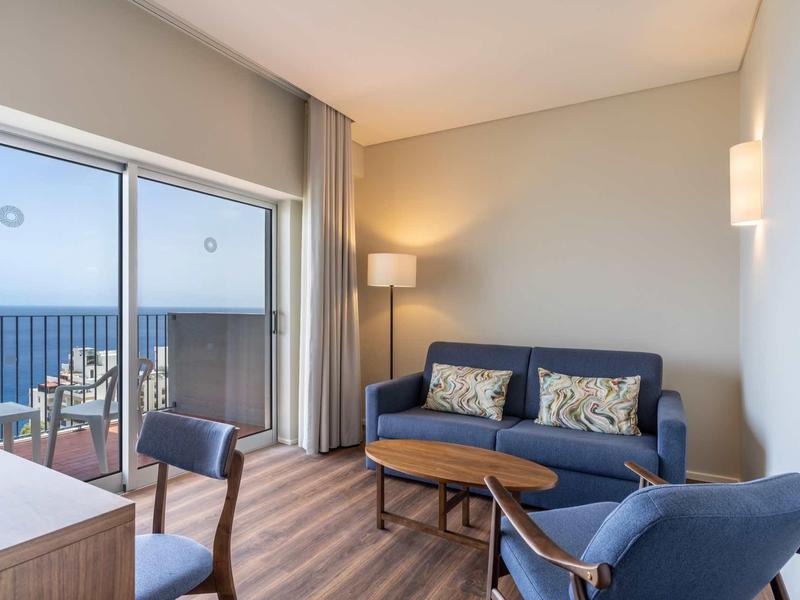 Gemütliches Wohnzimmer mit blauem Sofa, Sessel, Holztischen und Zugang zu Balkon mit Meerblick.