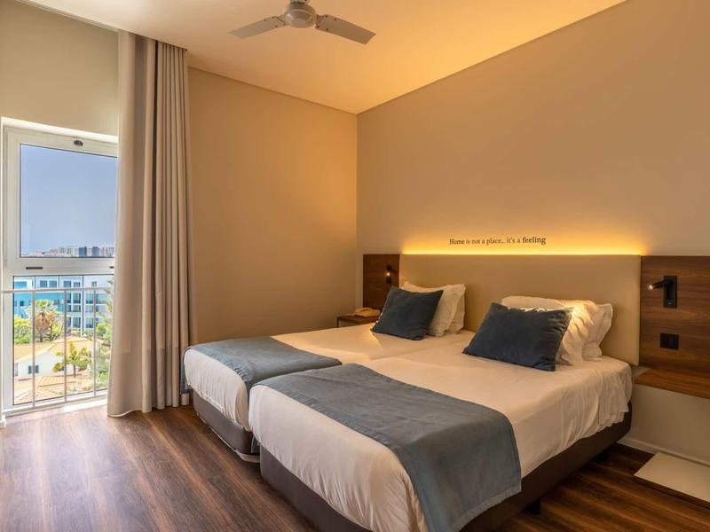 Modernes Doppelzimmer mit zwei Einzelbetten, Holzfußboden, Balkon und warmem LED-Licht hinter dem Kopfteil.